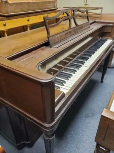 An old upright piano with damaged, missing, and uneven keys stands in a room with gray carpeting.