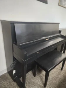 A black upright piano with a matching bench sits on a carpeted floor against a white wall.