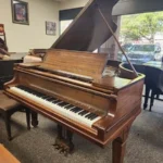 A brown grand piano with its lid open sits in a carpeted room with large windows and framed art on the walls.